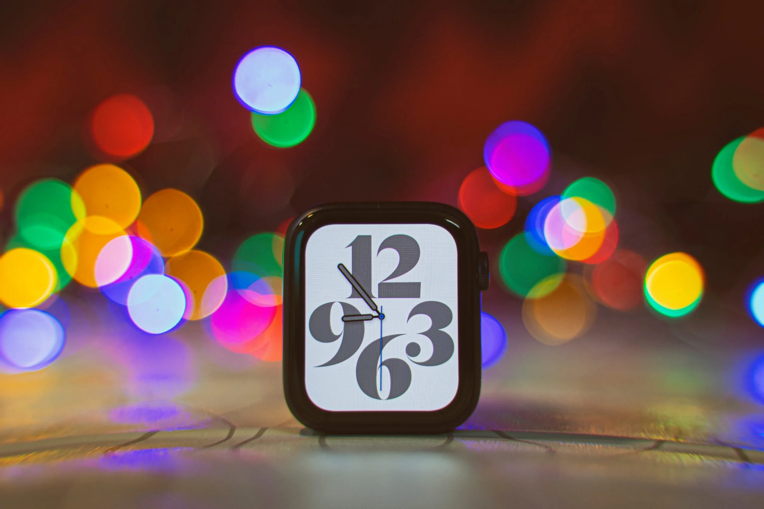 A photograph showing an Apple Watch Series 6 case sitting upright on a table, with a colorful lights in the background blurred with a depth-of-field effect A photograph showing an Apple Watch Series 6 case sitting upright on a table, with a colorful lights in the background blurred with a depth-of-field effect