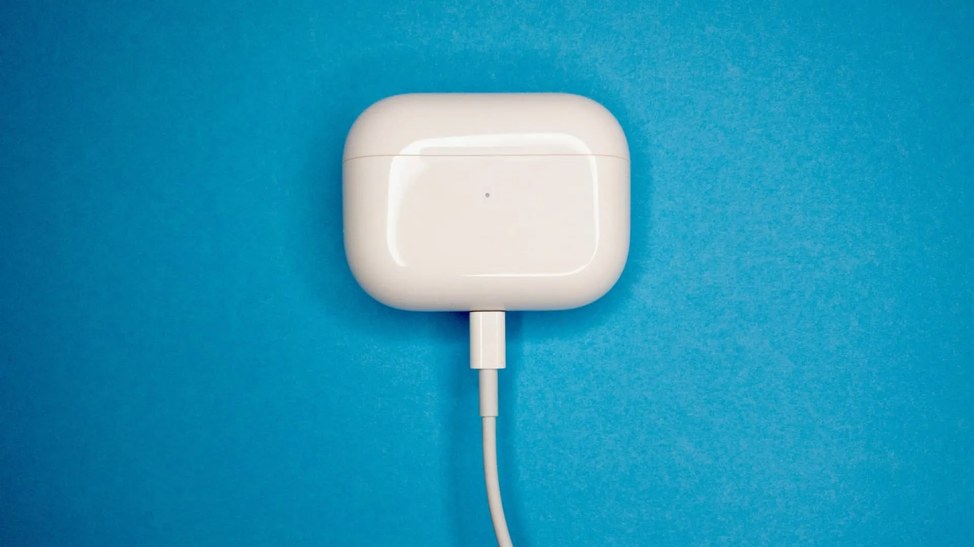 AirPods Pro kotelossaan, latautuu Lightning–USB-kaapelilla, asetettu sinistä teksturoitua taustaa vasten