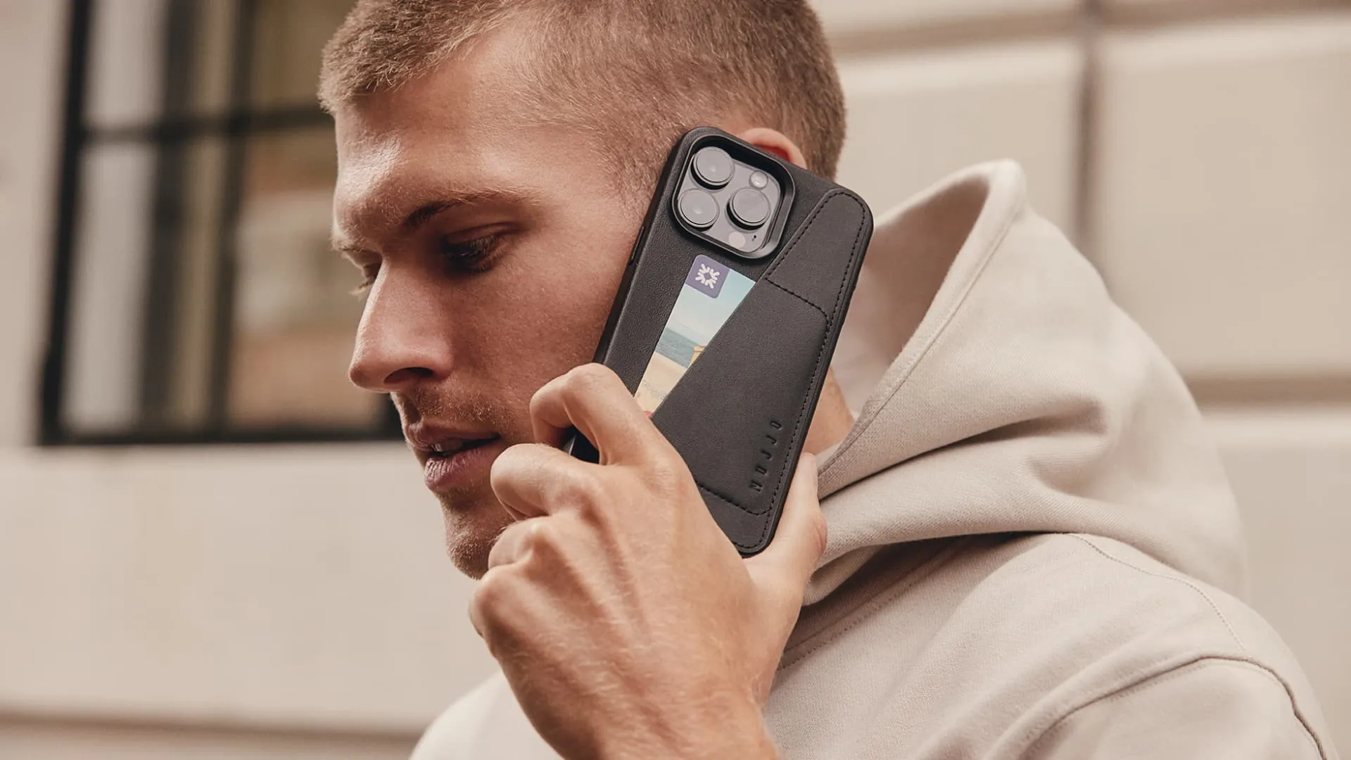 Vaikinas prie ausies laiko iPhone 15 Pro Max odiniame Mujjo piniginės dėkle