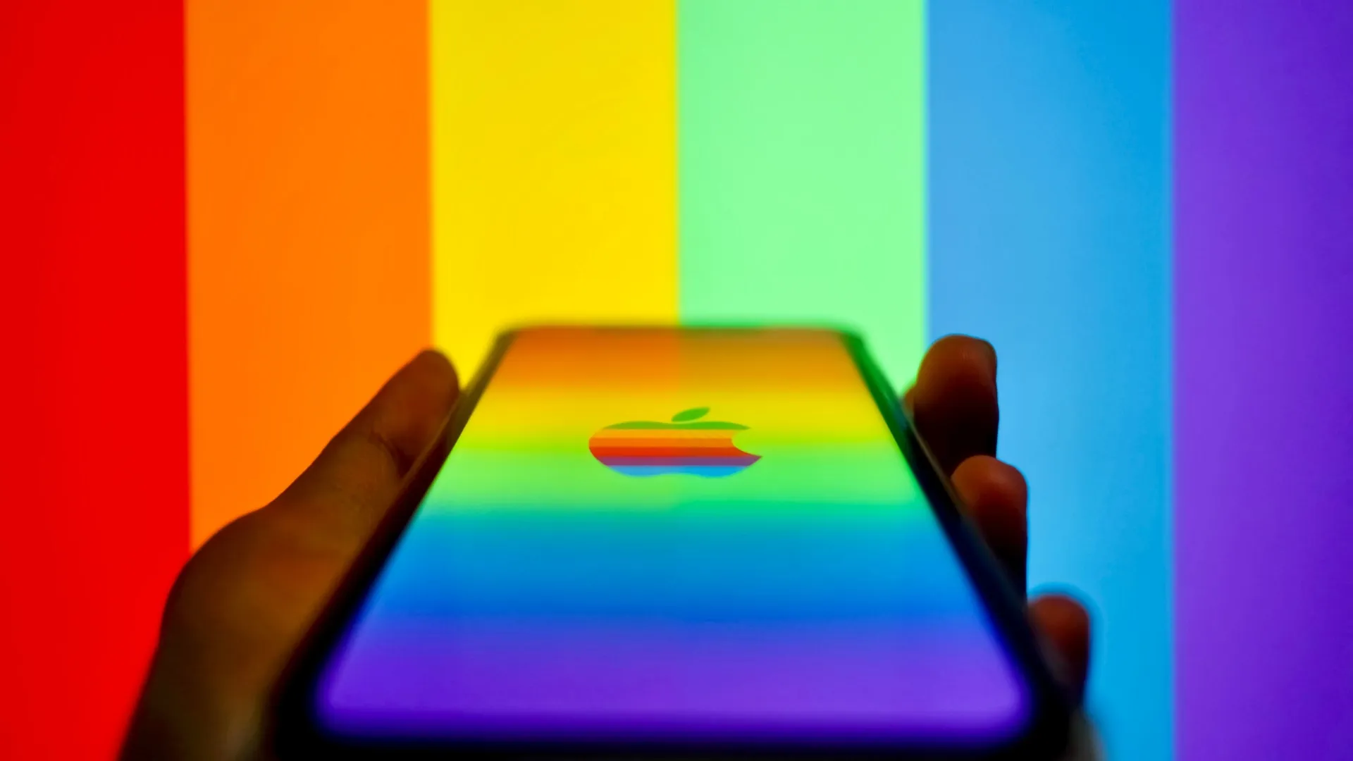 E segurando o iPhone exibindo um logotipo da Apple em arco-íris na tela, contra listras de arco-íris no fundo