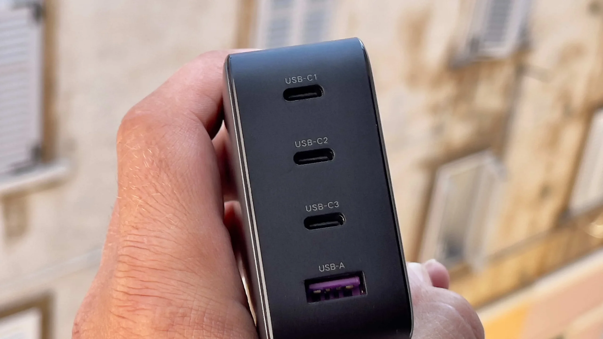 A male hand holding Ugreen's 100-watt Nexus power adapter, showcasing the charger's three USB-C ports and one USB-A port on the rear A male hand holding Ugreen's 100-watt Nexus power adapter, showcasing the charger's three USB-C ports and one USB-A port on the rear