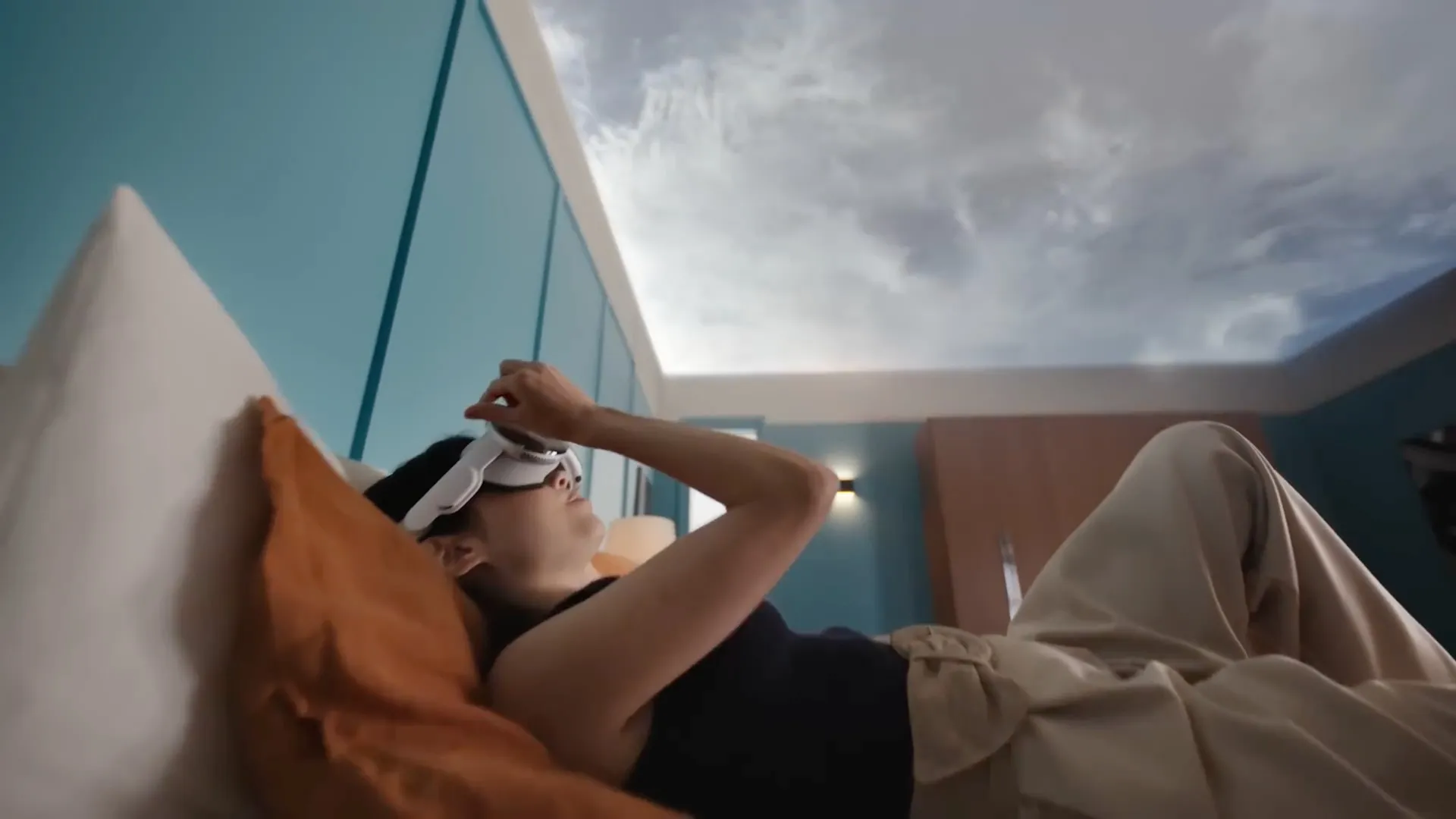 Ung kvinna som ligger på sängen med Apple Vision Pro-headsetet på sig, med utsikten över hennes tak ersatt av en virtuell himmelmiljö