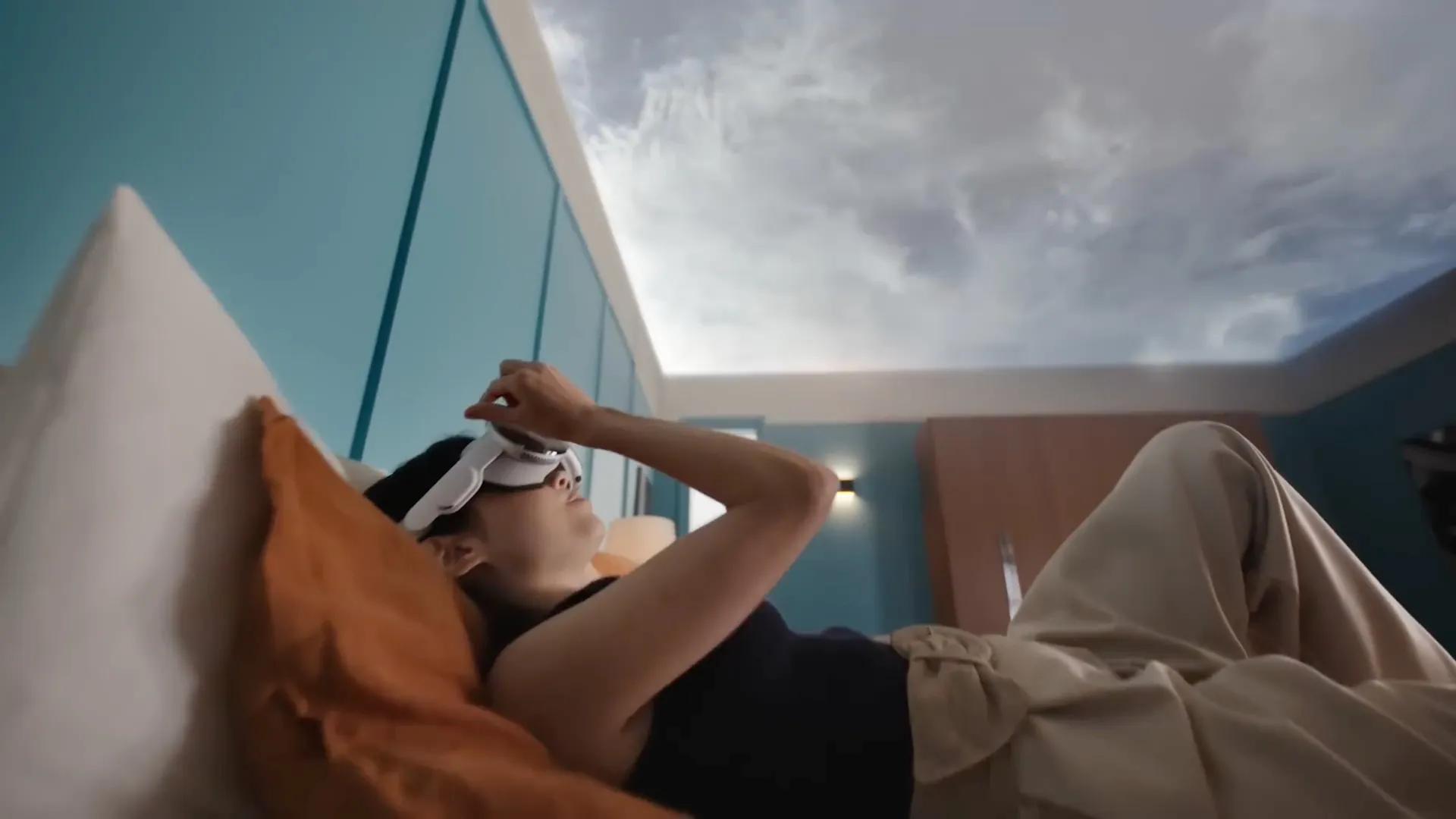 Jeune femme allongée sur son lit tout en portant un casque Apple Vision Pro, la vue de son plafond étant remplacée par un environnement de ciel virtuel